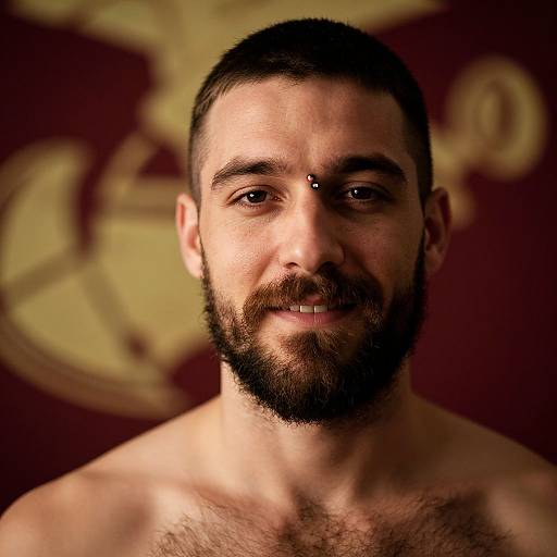 Photograph of a shirtless, bearded man with short dark hair, nose piercing, and chest hair, smiling against a blurred, warm-toned