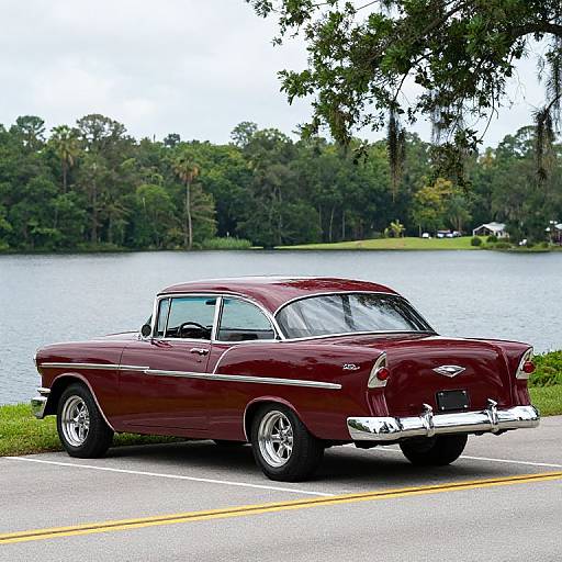 Photograph of a classic maroon 1950s Chevrolet sedan parked by a serene lakeside, with lush green trees in the background.