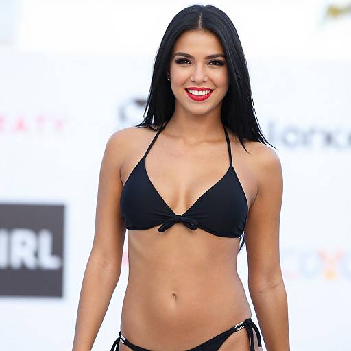 Photograph of a smiling young woman with long black hair, wearing a black bikini, standing against a bright, blurred outdoor background.