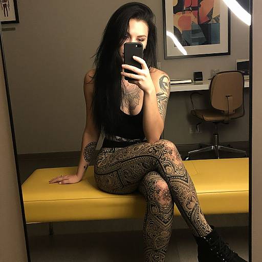 Photograph of a tattooed woman with long black hair, wearing a black top and intricate lace-patterned leggings, sitting on a yellow bench, taking