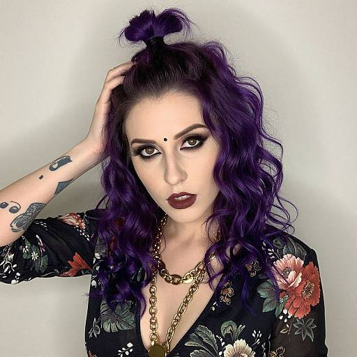 Photograph of a pale-skinned woman with purple curly hair, dark lipstick, floral black top, gold necklace, hand in hair, tattoos, white