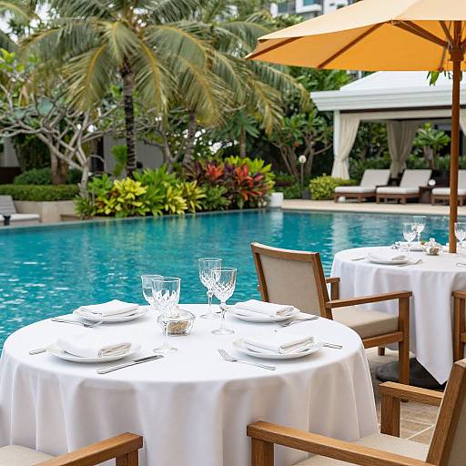 Photograph of a tropical outdoor poolside dining area with white-clothed round tables, crystal glasses, wooden chairs, orange umbrella, and lush green