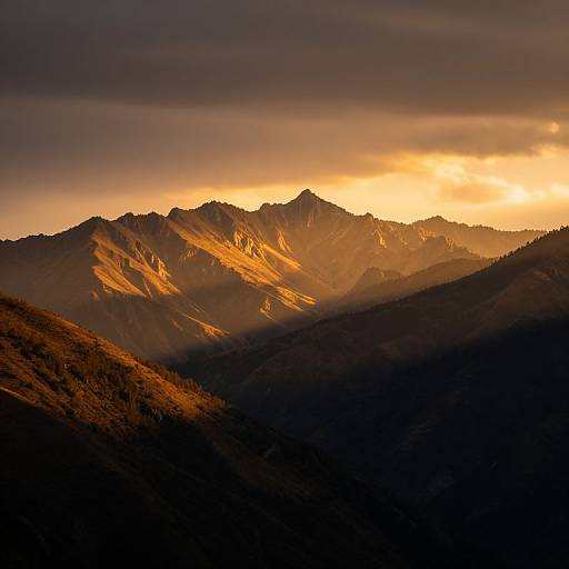 Photograph of a mountain range at sunset, with golden sunlight illuminating the peaks, casting long shadows, and a dark, cloudy sky overhead.