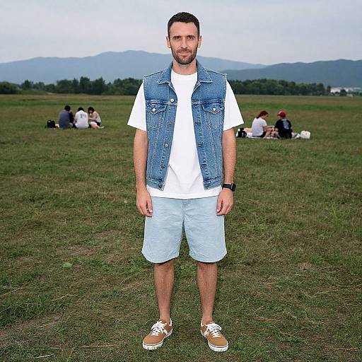 Photograph of a bearded man with short dark hair, wearing a denim vest, white t-shirt, light blue shorts, and brown sneakers, standing