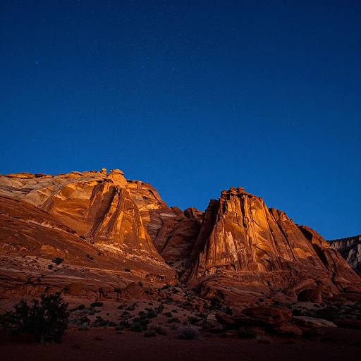 Photograph of a dramatic desert mountain range at sunset, with vibrant orange and red rock formations illuminated against a deep blue, starry night sky.
