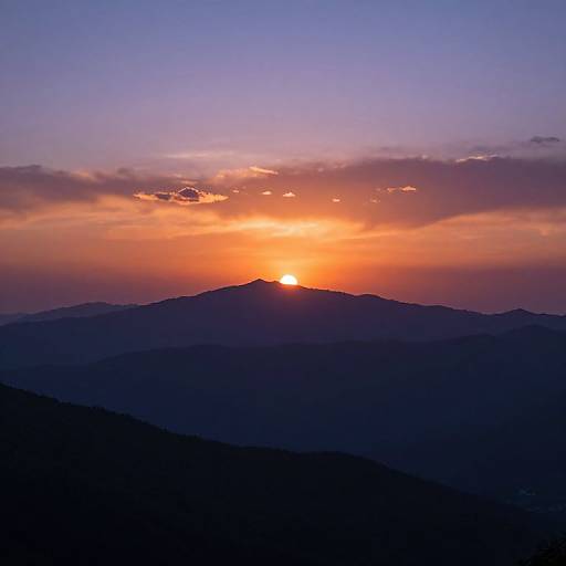 Photograph of a vibrant sunset over silhouetted mountain peaks, with the sun partially visible, casting an orange and purple sky.