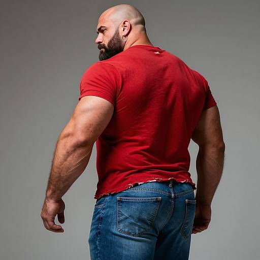 Photograph of a muscular, bald, bearded man in a red, slightly torn t-shirt and blue jeans, standing with his back to the camera