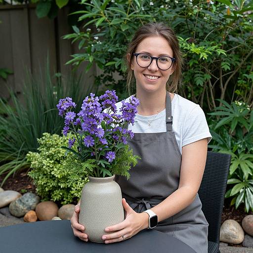 Photograph of a smiling woman with glasses, brown hair in a bun, white shirt, gray apron, holding a vase of purple flowers, seated