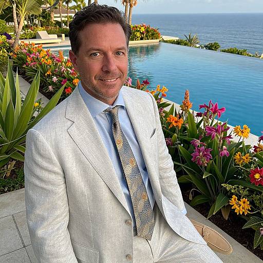 Photograph of a smiling middle-aged man in a light gray suit and patterned tie, seated by a vibrant, flower-lined pool overlooking the ocean.