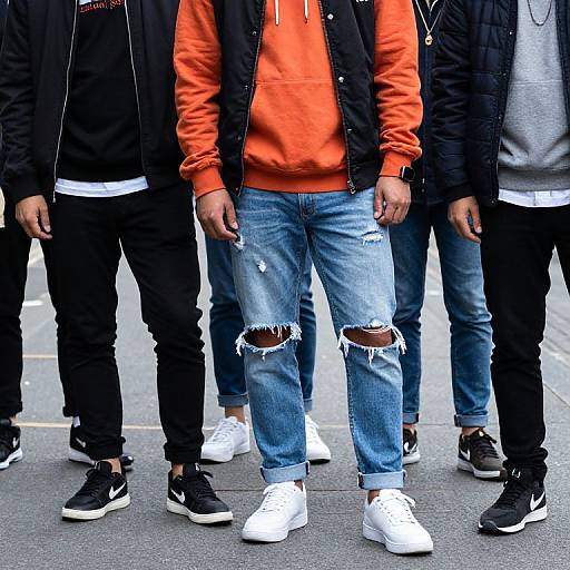 Photograph of four men, centered in orange hoodie with black vest and ripped blue jeans, standing in line, wearing white sneakers.