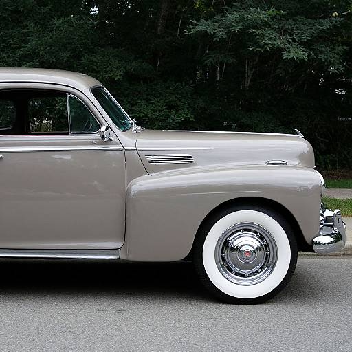 Photograph of a vintage silver car with white-walled tires, side profile, parked on a gray road, against a dark green forest background.