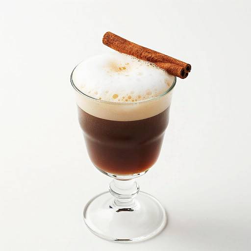 Photograph of a glass cocktail with dark brown liquid, topped with a cream layer and a cinnamon stick garnish, on a white background.