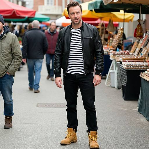 Photograph of a stylish man in a black leather jacket, striped shirt, black pants, and tan boots, standing in a bustling outdoor market with colorful