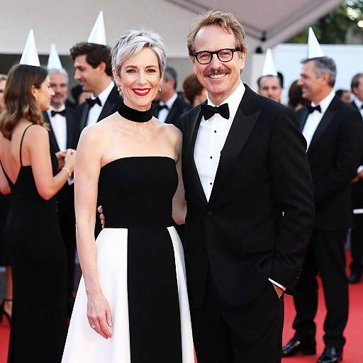 Photograph of a red carpet event featuring a smiling woman with short white hair in a black-and-white strapless dress, and a man with glasses and