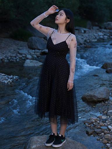 Asian woman with tattoos in sheer-sleeved, black, tulle dress and black sneakers, standing by a rocky, blue-lit stream.
