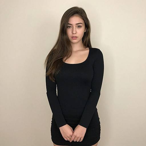Photograph of a young woman with long dark brown hair, wearing a black long-sleeve dress, standing against a plain white background, looking directly