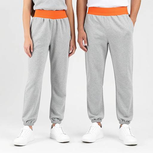 Photograph of two people from waist to ankles, wearing gray sweatpants with orange waistbands, white socks, and white shoes, standing against a white