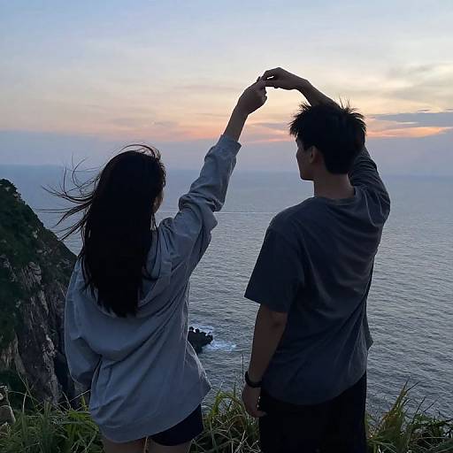 Photograph of a couple standing on a cliff at sunset, silhouetted against the ocean, with the woman raising her hand and tousled hair