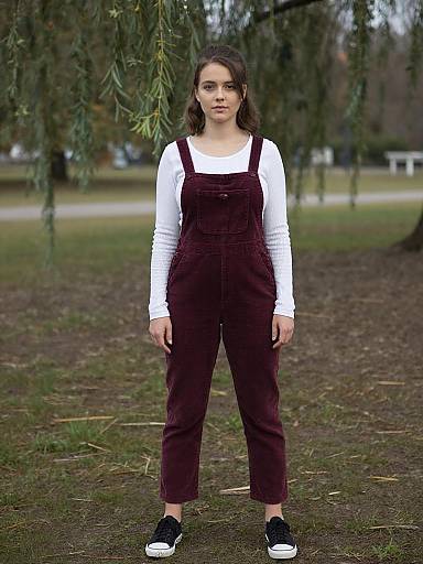 Photograph of a young woman with long brown hair, wearing a white long-sleeve shirt and maroon overalls, standing outdoors on grass with