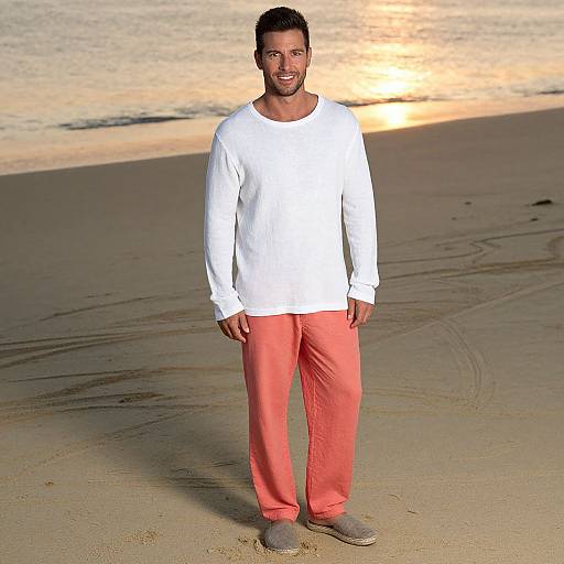Photograph of a smiling Black man with short hair, wearing a white long-sleeve shirt, coral pants, and gray sneakers, standing on a