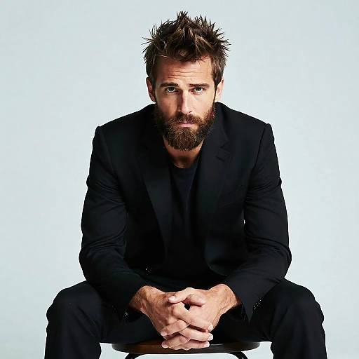 Photograph of a rugged, bearded man with spiky brown hair, wearing a black suit, sitting with hands clasped, intense gaze, white