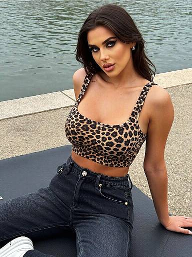 Photograph of a brunette woman with medium skin tone, wearing a leopard-print crop top and high-waisted black jeans, sitting on a black mat