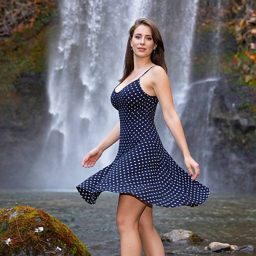 Photograph of a young woman with light brown hair in a navy blue, polka-dot dress, standing in front of a waterfall, holding her skirt