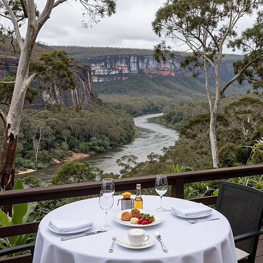 Photograph of a round table with white tablecloth and meal setup, overlooking a lush river valley and towering cliffs in a forested area.