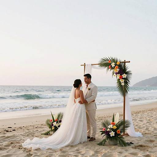 Photograph of a bride in a white gown and groom in a beige suit, standing under a floral arch on a beach.