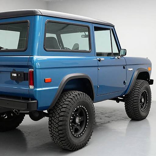 Photograph of a blue Jeep Wrangler with black off-road tires and black rims, viewed from the rear left in a bright, white-walled garage