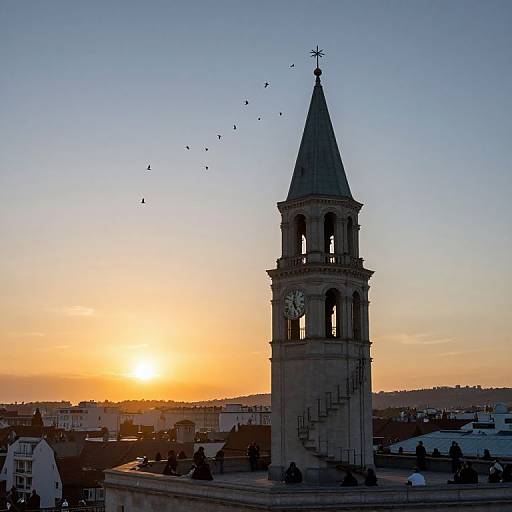 Photograph of a sunsetting sky with a tall, stone clock tower, birds flying, and urban buildings below, casting warm orange and blue hues.