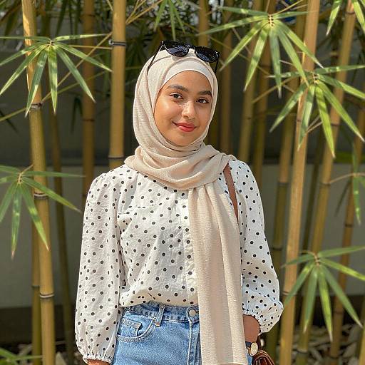Photograph of a smiling young woman with light brown skin, wearing a white polka-dot blouse, beige hijab, and blue jeans, standing in