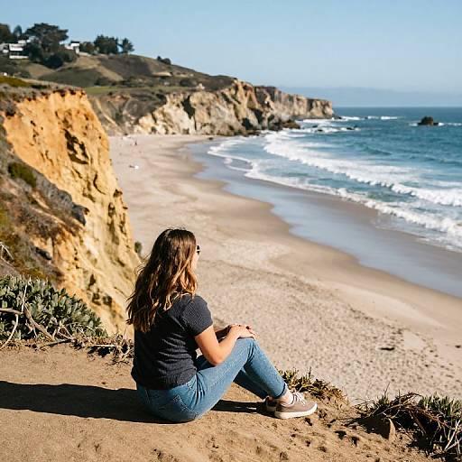 Photograph of a woman with wavy brown hair, wearing a black t-shirt and blue jeans, sitting on a sandy cliff overlooking a sandy beach with