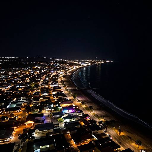 Aerial nighttime photograph of a brightly lit coastal city, showcasing a glowing grid of lights, dark ocean, and illuminated streets.