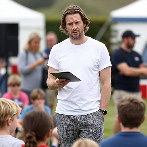 Photograph of a middle-aged man with shoulder-length brown hair, white t-shirt, and grey patterned pants, holding a tablet, speaking to a
