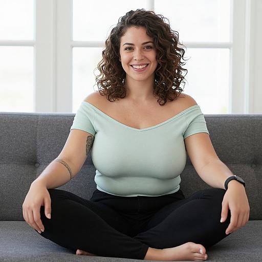 Photograph of a smiling, curly-haired woman with light skin, wearing a light blue off-shoulder top and black pants, sitting cross-legged on