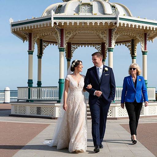 Photograph of a bride in a white lace gown, groom in a black suit, and a woman in a blue blazer walking under a Victorian-style