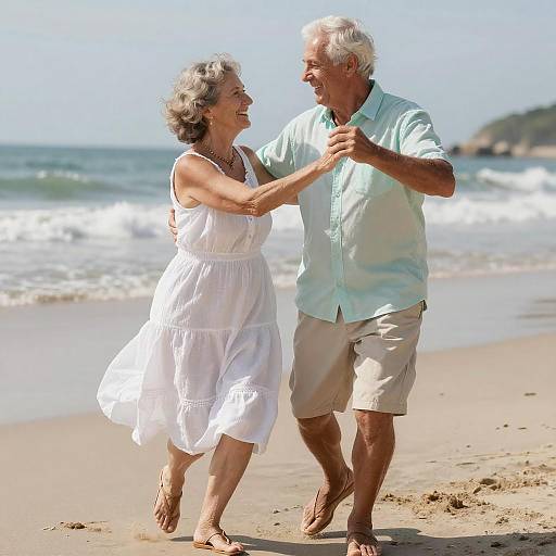Photograph of elderly couple dancing joyfully on sunny beach; woman in white dress, man in light shirt and beige shorts, waves in background.