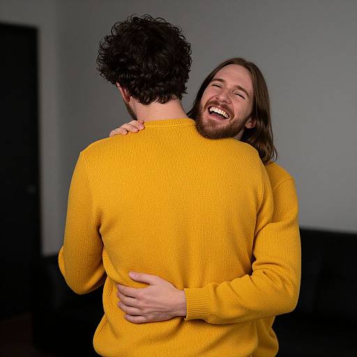 Photograph of two men hugging; one with curly black hair, yellow sweater, facing away; other with long brown hair, beard, smiling,