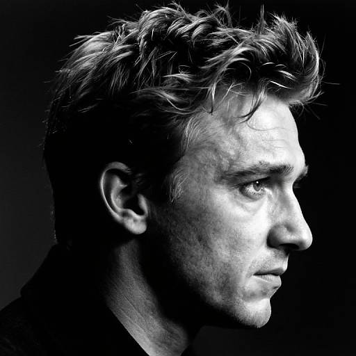 High-contrast black-and-white portrait of a serious, profile-view young man with tousled hair, sharp features, and intense gaze against a dark background
