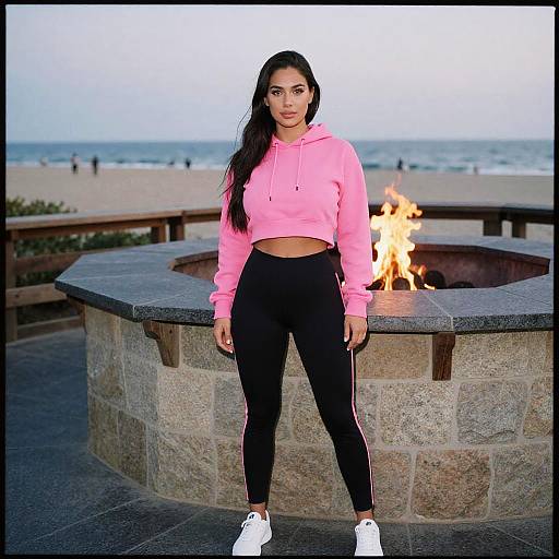 Photograph of a young woman with long black hair, pink hoodie, black leggings, white sneakers, standing in front of a stone fire pit on a