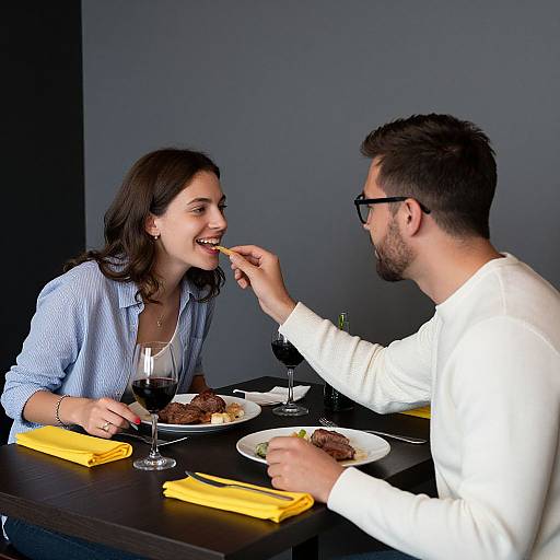 Photograph of a smiling woman and bearded man at a dark wooden table, feeding her a bite of meat while dining together. Both wear casual clothes