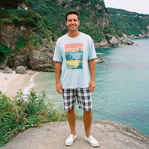 Photograph of a smiling man with short dark hair, light blue t-shirt, plaid shorts, and white sneakers, standing on a cliff overlooking a