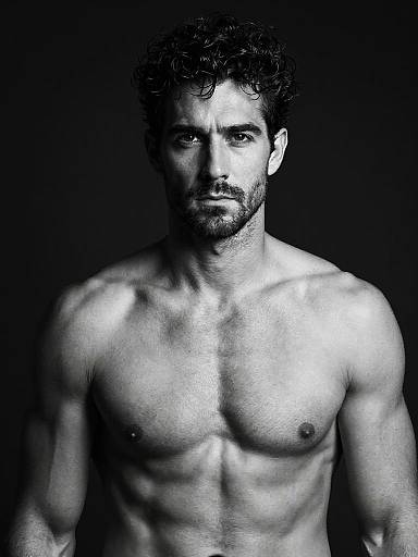 Black-and-white photograph of a muscular, shirtless man with curly hair and beard, staring intently at the camera against a dark background.