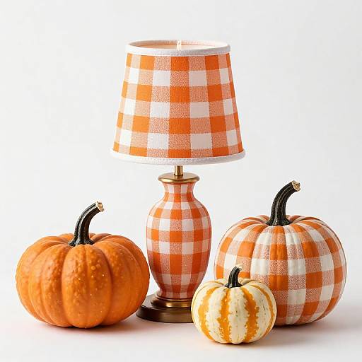 Photograph of three pumpkins and a lamp with orange-and-white checkered fabric, standing on a white background. Lamp has a gold base.