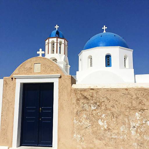 Photograph of a Greek island church with white walls, blue domes, black door, cross accents, and clear blue sky.