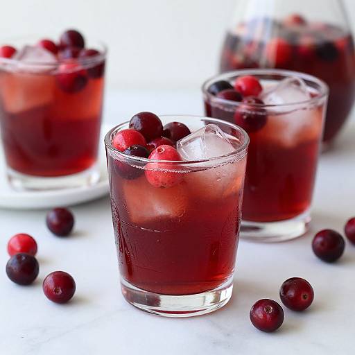 Photograph of three glass cups filled with dark red cranberry juice, ice cubes, and fresh cranberries, with scattered berries on a white surface.