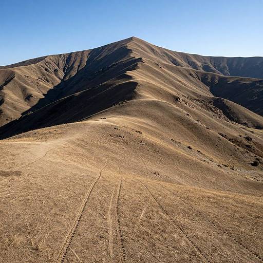 Photograph of a sunlit, barren, brown mountain with visible tire tracks, casting deep shadows, under a clear, bright blue sky.