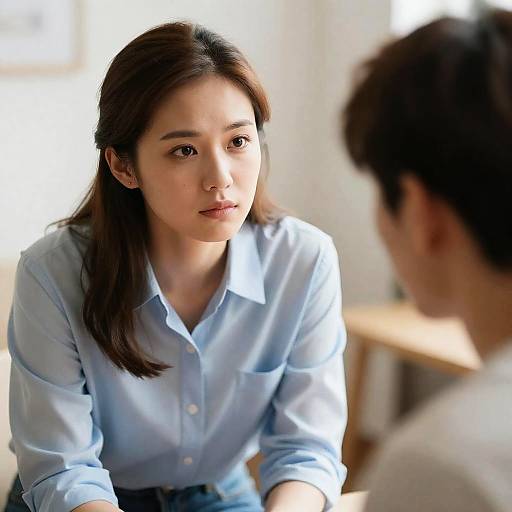 Photograph of an Asian woman with straight black hair, wearing a light blue button-up shirt, intently looking at a man with short brown hair,