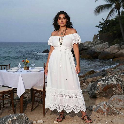 Photograph of a curvy woman with dark curly hair wearing a white off-shoulder lace dress, standing by a beach table with a floral centerpiece